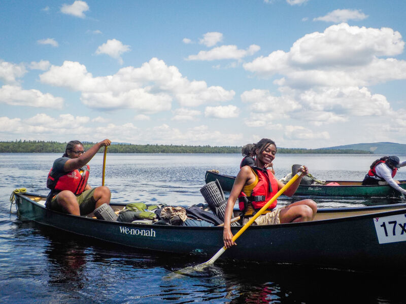 The image shows a group of people canoeing on a lake. The sky is blue with some clouds. The people are wearing life jackets and appear to be enjoying themselves. The canoes are moving through the water. The overall scene is peaceful and suggests a recreational activity.